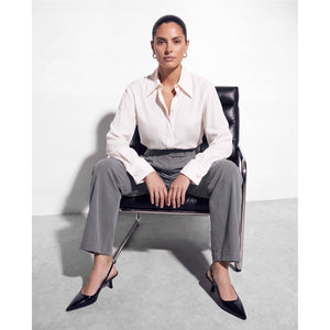 Model seated facing forward in a black leather chair, wearing a Femponiq ivory shirt with an open pointed collar and wide buttoned cuffs, grey tailored trousers and black pointed-toe slingback heels.