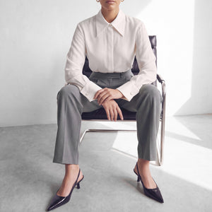 A model sits on a modern metal chair wearing a white long-sleeve collared blouse, gray wide-leg trousers, and black pointed-toe heels in a bright minimalist room.
